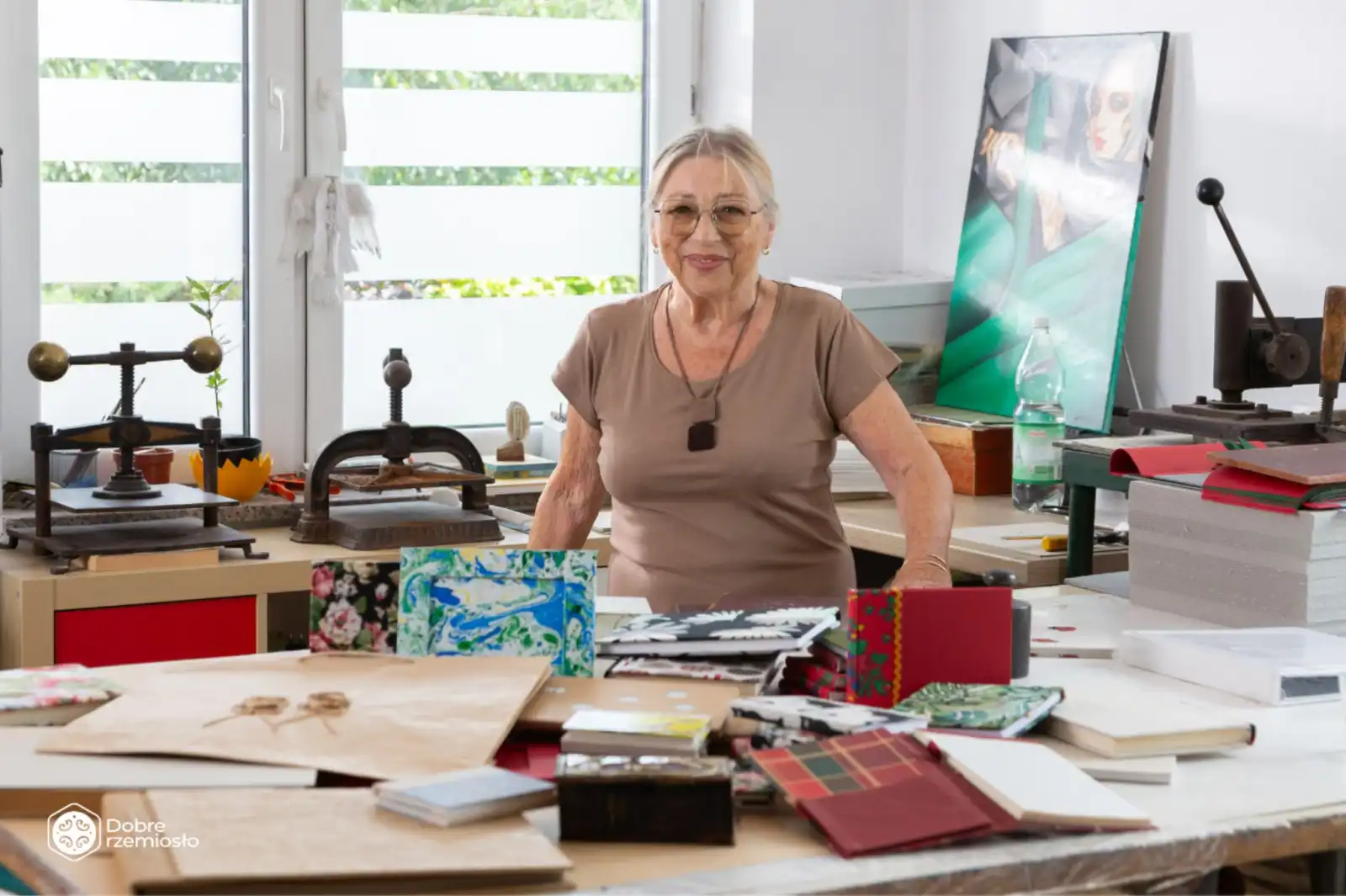 Na pierwszym tle jest stół z albumami i książkami wykonanymi własnoręcznie przez introligatorkę. Za stołem stoi Nina Teresa Radkiewicz - jasnowłosa, uśmiechnięta osiemdziesięcioletnia pani.